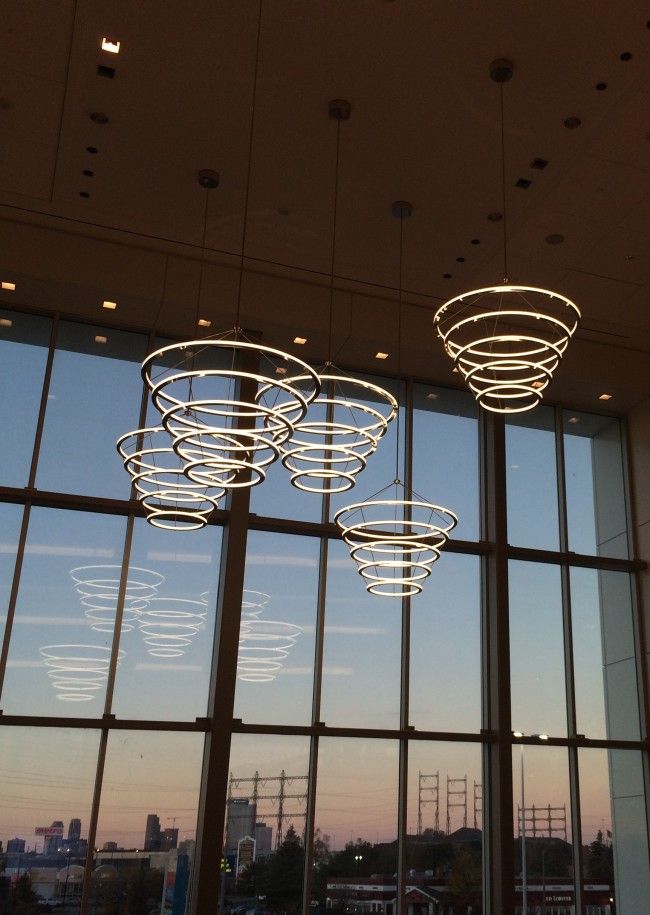 A low-angle photo of Sherway Gardens' custom Halo chandeliers from Roll & Hill. Through the glass wall, twilight falls.