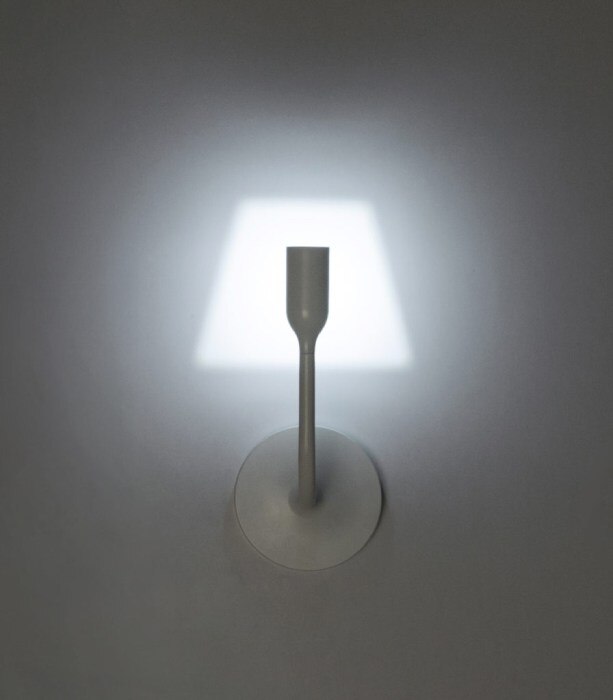 YOYWall wall sconce, designed for Innermost by YOY Studio, casts its inimitable lampshade-shaped light upon a wall, seen here from the front in a darkened room.