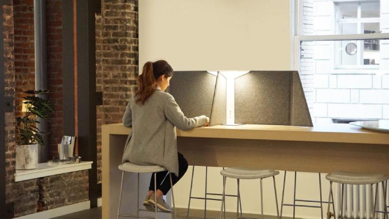 A woman works in an open-concept office, shielded and illuminated by Corner Office 30 from Pablo Design.