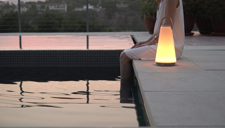 A woman sits, dipping her feet in a backyard pool, with UMA Sound Lantern portable table light at her side on the deck.