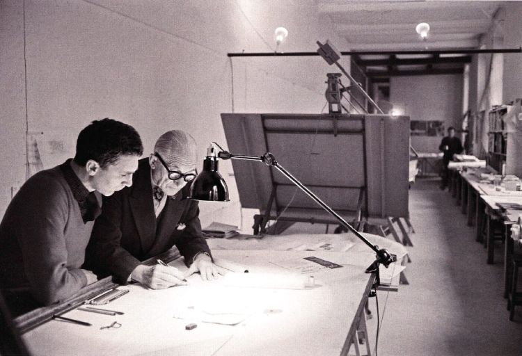 An aging Le Corbusier works at a drafting table, under the light of a Lampe Gras, with an assistant in his atelier in 1959