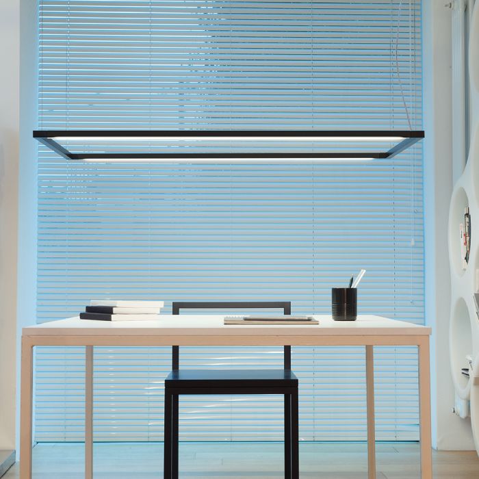 Spigolo Horizontal suspension light illuminates a simple table desk next to a circle-based shelving unit in a minimalist office decorated in a simple black-and-white colour scheme.