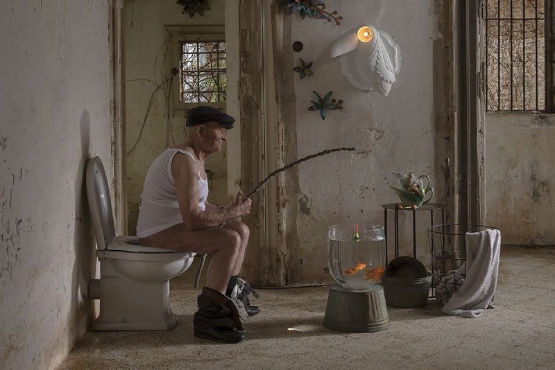 An aging man in a peaked cap sits upon a toilet and fishes in a goldfish bowl under the light of Cubano wall light from Karman, in one of the company's distinctively eccentric photos.