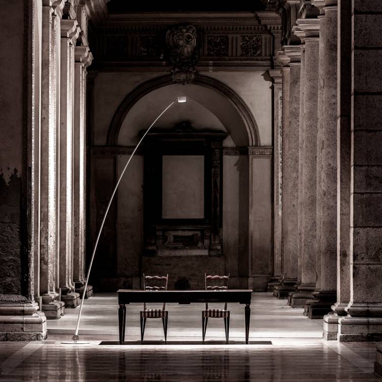 Sampei floor light illuminates a wooden table and chairs in the middle of a vast baroque hall.