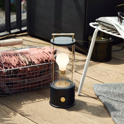 The Muse lantern by Tala sits on a wooden deck with a basket, a chair, and a watering can.