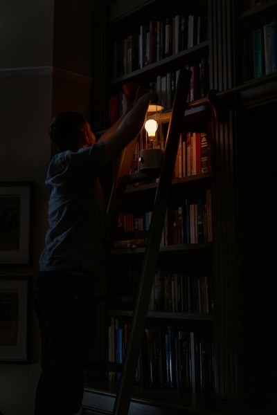 A dark figure stands on a ladder searching for a book in a tall bookshelf by the light of Tala's The Muse lantern.