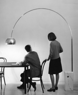 A vintage marketing photo of Arco floor light from Flos. A man sits at a table with a woman behind him, both facing away from camera. Arco illuminates the table on which he works.