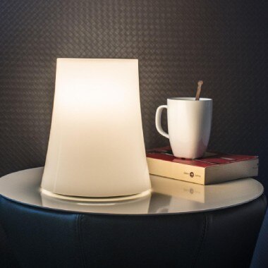 Birdie Zero table light, designed by Ludovica and Roberto Palomba for Foscarini, sits on a side table next to a book, on top of which sits a coffee mug with a spoon in it.