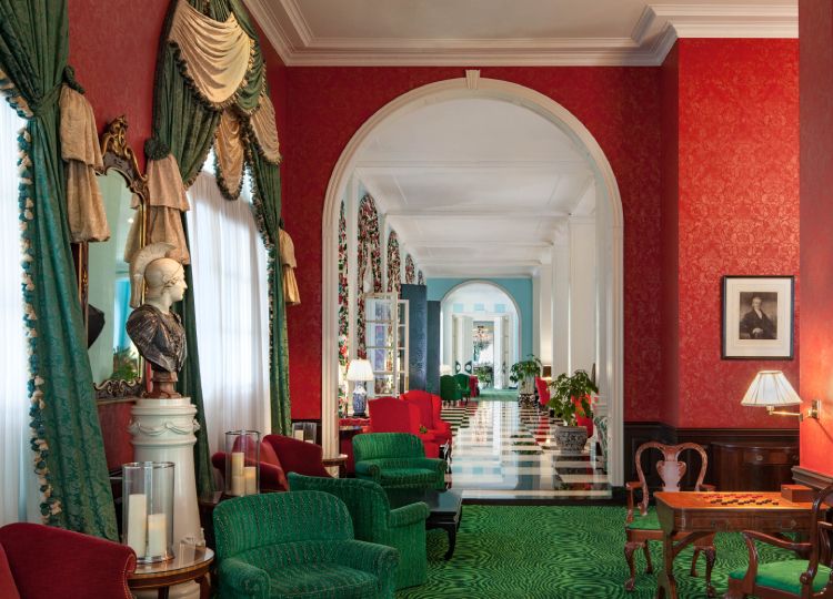 A hallway in the Greenbrier Hotel, viewed through an archway from the sitting room. Bright red and green colours contrast with wood accents. The hall floor is a checkered black and white. A bust of a helmeted Athenian warrior (possibly Athena herself) sits on a pedestal between two windows with Baroque curtains on the left of frame.