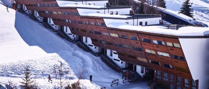 A wide shot of La Cascade residence at Les Arcs ski resort. A reddish-brown and white building, it suggests a series of stairs placed on the hillside, which it follows along the incline.