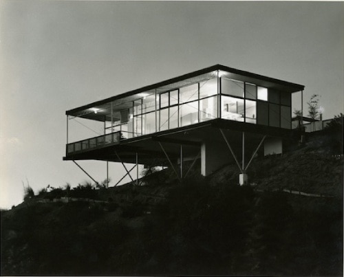 A black-and-white photo of a mid-century modern architectural residence on a hill in California. It hangs out from the hillside, supported from below by a few well-placed beams.