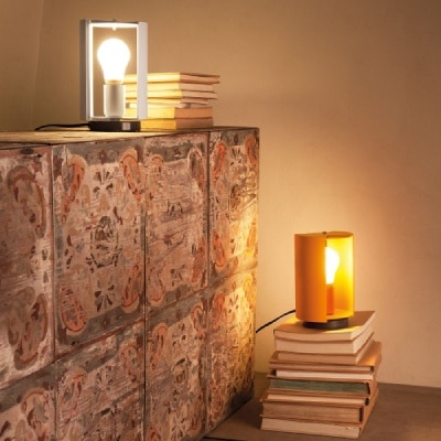 A pair of Pivotante À Poser table lamps. The yellow one sits on a stack of books next to a counter of ceramic tile, while the white sits upon the counter next to its own stack of books.