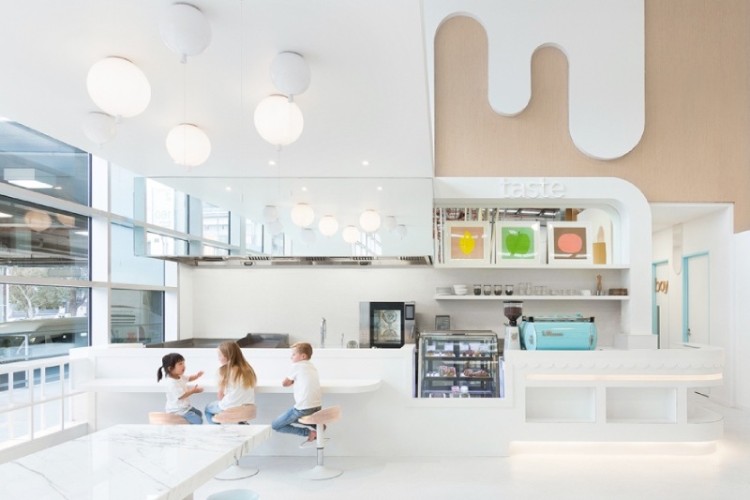 A bright pastel-coloured café illuminated by a cluster of Memory ceiling lights. A trio of children sit at the counter next to a glass case of delectable baked goods.