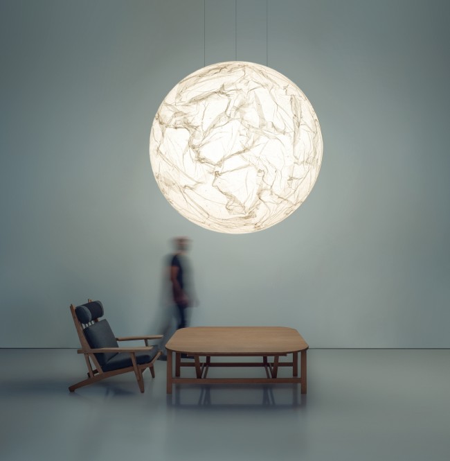 Moon 200 suspension light, designed and released by Davide Groppi, hangs over a table with a blurred figure in the background in a studio photo.