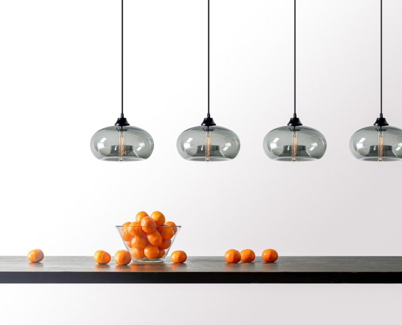 A linear cluster of Aurora pendant lights hang over a black counter with a bowl of oranges on a minimalist studio set.