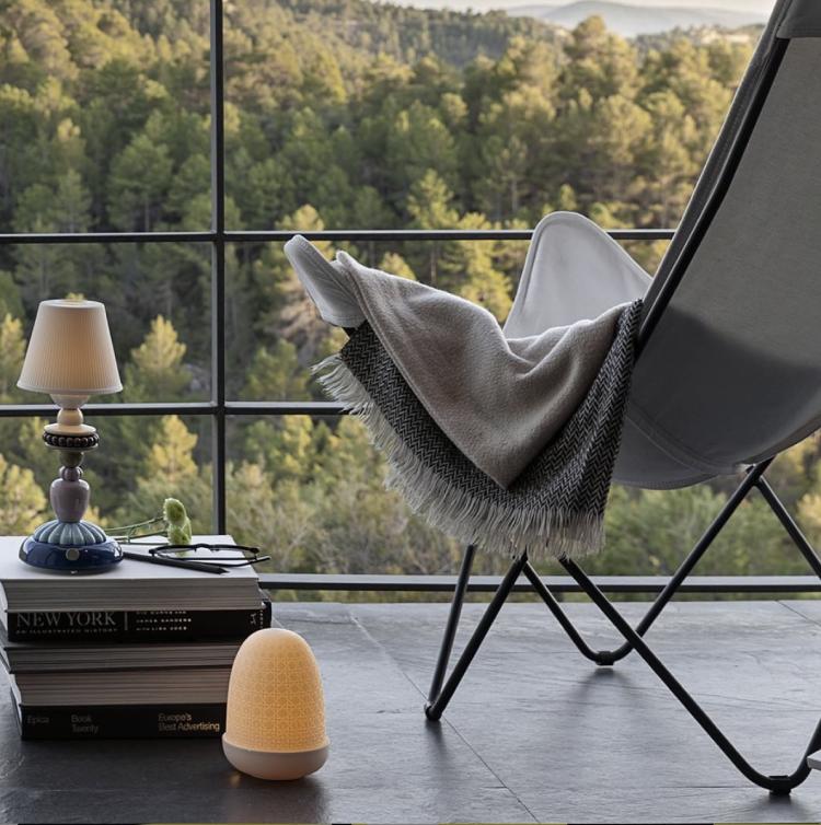 Lladro portable table lamps Wicker Dome and Sunflower Firefly sit beside a contemporary chair overlooking woods.