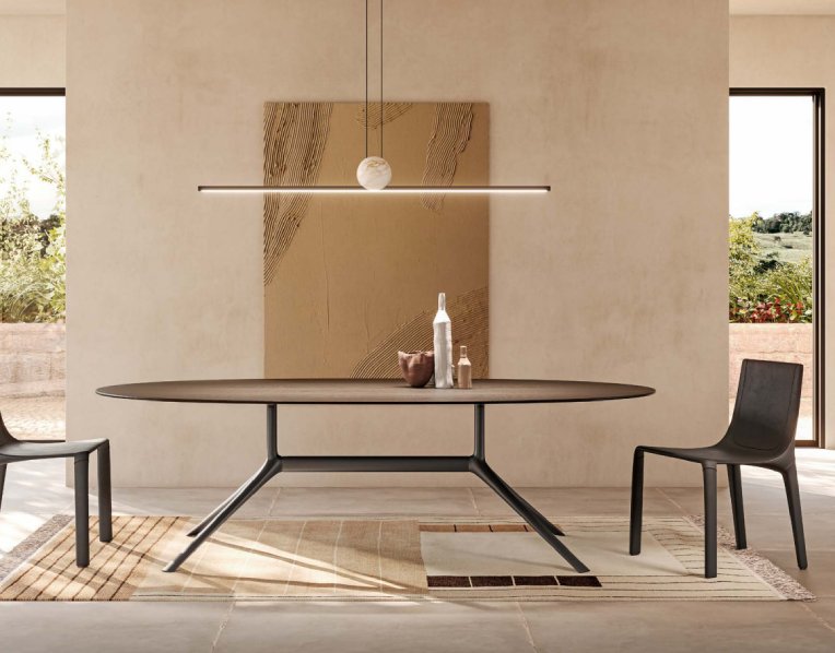 A warm and airy dining room decorated in earth tones. Over the oval dining room table hangs an illuminated Regolo suspension light, designed by Stefano Traverso for Italamp. Through patio doors on either side of the image, a lush countryside can be seen.