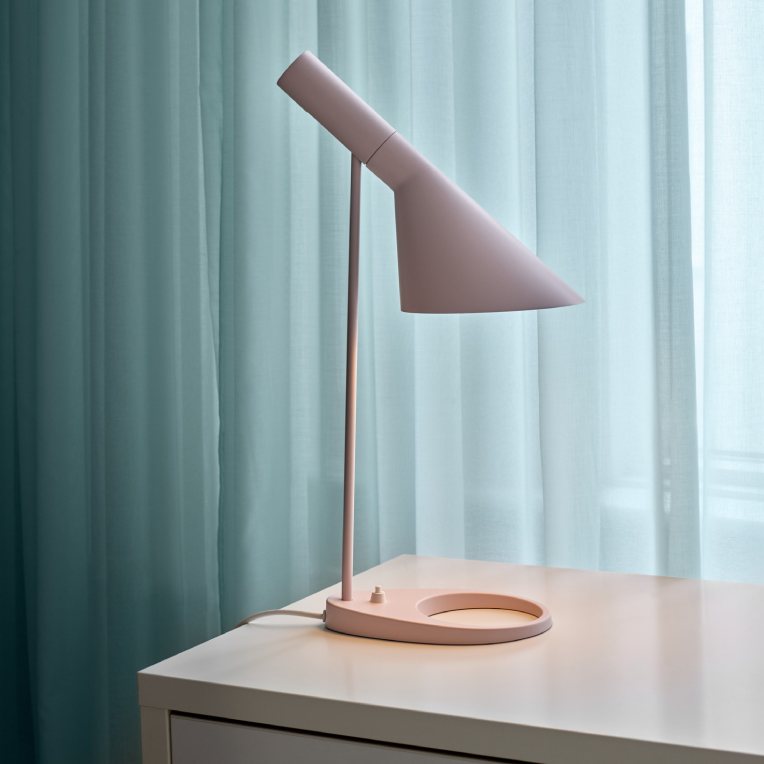 Arne Jacobsen's AJ table lamp sits on a wooden dresser in a bright modern bedroom. Sunlight filters through diaphanous curtains behind.