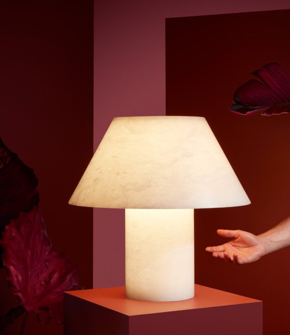 Petra M GR table light, designed for Spanish lighting brand Parachilna by Jordi Veciana, sits on a red-toned studio set. A hand reaching in from out of frame catches Petra's light in its palm.