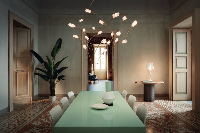 A spacious, comfortable dining room. Over the long, green dining table hangs Slamp's new Tulip cluster suspension light, designed by Marc Sadler. Through an open doorway in the centre of the dining room wall, a darkened room is visible, illuminated by sunlight filtering through a tall window.