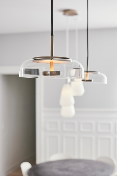 A pair of Nuura's Blossi pendants in Nordic Gold with transparent glass diffusers hang above a kitchen counter. Out of focus in the background, a Anoli triple cluster suspension light can be seen above a dining table.