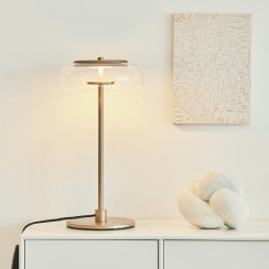 Blossi table lamp in Nordic Gold with a transparent glass diffuser sitting on a white credenza with a decorative object