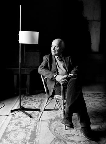 Miguel Milá sitting on one of his designs, the Salvador Chair, next to his TMC Floor Lamp